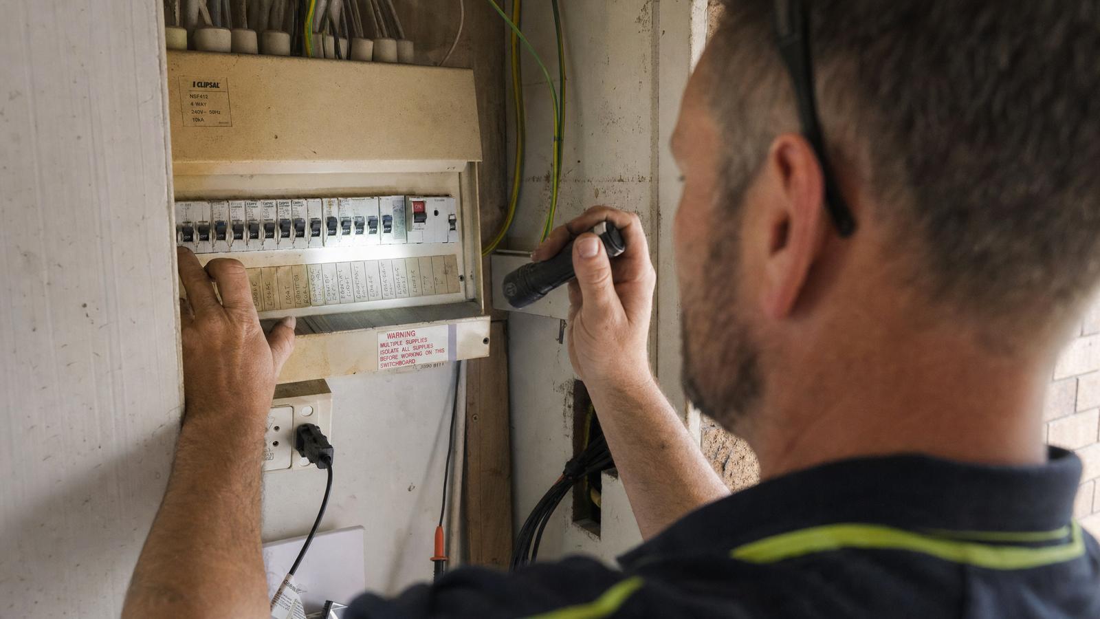 Electrician inspecting an older residential electrical switchboard with a torch before a battery installation